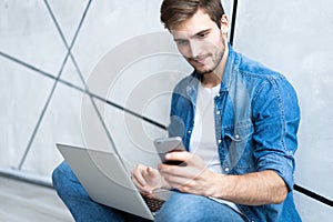 Young caucasian man sitting over gray wall using computer laptop and smartphone.