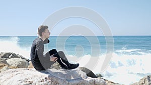 Young caucasian man sitting on the rocks with fingers in his hair. Strong ocean waves hitting the rocky beach with water splashing