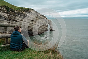 Young caucasian man sitting on top of the cliff and looking at Cape Farewell rock formation. New Zealand