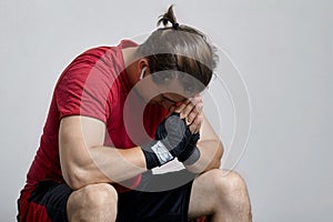 Young caucasian man in sport wear and boxing bandages sits with hands to forehead. Pray, or despair moment, sorrow, hopelessness,