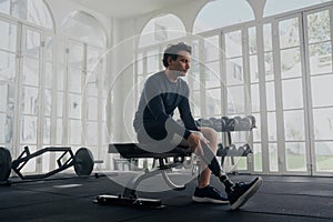 Young caucasian man in sports clothing sitting on bench press while attaching prosthetic leg at the gym