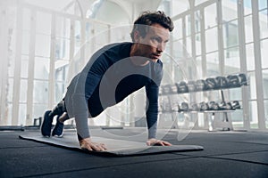 Young caucasian man in sportswear in plank position while doing push-ups on exercise mat at gym