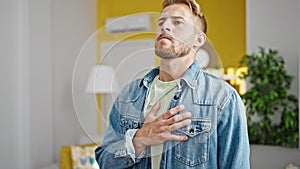 Young caucasian man standing with hand on heart listening to anthem at home