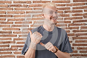 Young caucasian man standing over bricks wall pointing to the back behind with hand and thumbs up, smiling confident