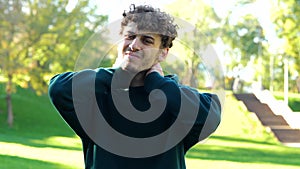 Young Caucasian man stretching before outdoor activities. Guy feeling pain in his neck standing in green park on sunny