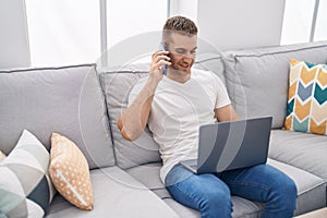 Young caucasian man talking on smartphone using laptop at home