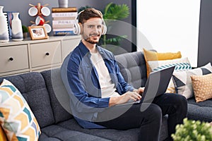 Young caucasian man using laptop and headphones sitting on sofa at home