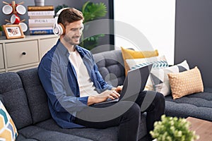 Young caucasian man using laptop and headphones sitting on sofa at home