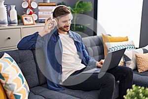 Young caucasian man using laptop and headphones sitting on sofa at home