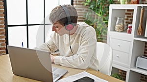 Young caucasian man using laptop and headphones writing on notebook at dinning room