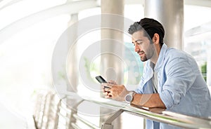 Young caucasian man using smart phone and smiling while waiting his friend at the outside. The man holding and using cellphone for