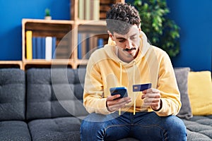 Young caucasian man using smartphone and credit card sitting on sofa at home