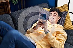 Young caucasian man using smartphone lying on sofa at home
