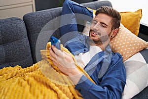 Young caucasian man using smartphone lying on sofa at home