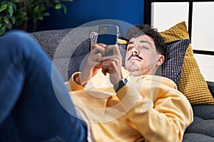 Young caucasian man using smartphone lying on sofa at home