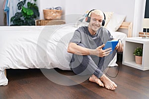 Young caucasian man using touchpad and headphones sitting on floor at bedroom