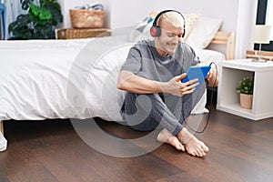 Young caucasian man using touchpad and headphones sitting on floor at bedroom