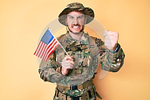 Young caucasian man wearing camouflage army uniform holding usa flag annoyed and frustrated shouting with anger, yelling crazy