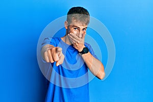 Young caucasian man wearing casual blue t shirt laughing at you, pointing finger to the camera with hand over mouth, shame