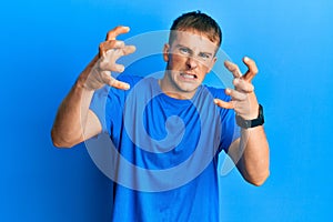 Young caucasian man wearing casual blue t shirt shouting frustrated with rage, hands trying to strangle, yelling mad
