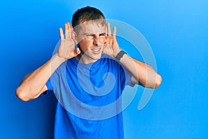 Young caucasian man wearing casual blue t shirt trying to hear both hands on ear gesture, curious for gossip