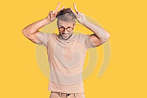 Young caucasian man wearing casual clothes and glasses posing funny and crazy with fingers on head as bunny ears, smiling cheerful