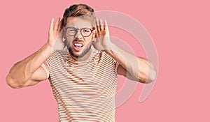 Young caucasian man wearing casual clothes and glasses trying to hear both hands on ear gesture, curious for gossip