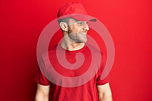 Young caucasian man wearing delivery uniform and cap looking away to side with smile on face, natural expression