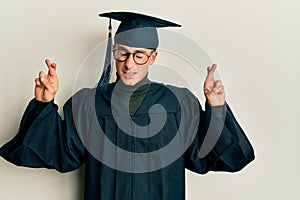 Young caucasian man wearing graduation cap and ceremony robe gesturing finger crossed smiling with hope and eyes closed