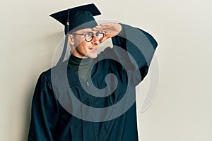 Young caucasian man wearing graduation cap and ceremony robe very happy and smiling looking far away with hand over head