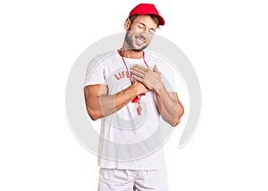 Young caucasian man wearing lifeguard t shirt holding whistle smiling with hands on chest with closed eyes and grateful gesture on