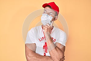 Young caucasian man wearing lifeguard t shirt using medical mask serious face thinking about question with hand on chin,