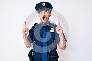 Young caucasian man wearing police uniform amazed and surprised looking up and pointing with fingers and raised arms