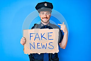 Young caucasian man wearing police uniform holding fake news banner pointing finger to one self smiling happy and proud