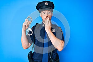 Young caucasian man wearing police uniform holding handcuffs covering mouth with hand, shocked and afraid for mistake