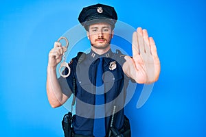 Young caucasian man wearing police uniform holding handcuffs with open hand doing stop sign with serious and confident expression,