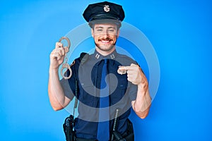 Young caucasian man wearing police uniform holding handcuffs pointing finger to one self smiling happy and proud