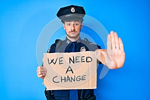 Young caucasian man wearing police uniform holding we need a change banner with open hand doing stop sign with serious and