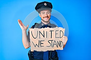 Young caucasian man wearing police uniform holding united we stand banner celebrating victory with happy smile and winner