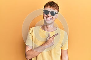 Young caucasian man wearing stylish sunglasses smiling cheerful pointing with hand and finger up to the side