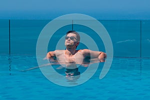 Young caucasian man wearing sunglasses at the edge of an infinity pool with blue sea and sky in the background