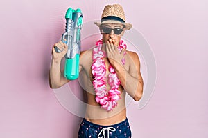 Young caucasian man wearing swimsuit and hawaiian lei holding watergun covering mouth with hand, shocked and afraid for mistake