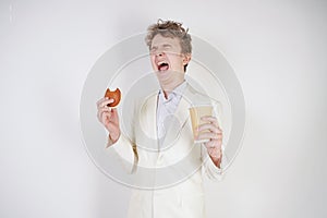 Young caucasian man in a white business suit stands with a paper Cup of coffee and a bun in his hand and suffers from severe abdom