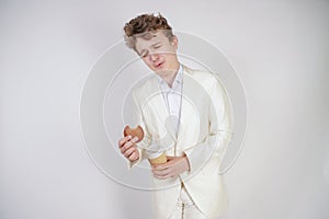 Young caucasian man in a white business suit stands with a paper Cup of coffee and a bun in his hand and suffers from severe abdom