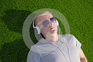 A young Caucasian man in a white T-shirt and headphones lies on an artificial lawn