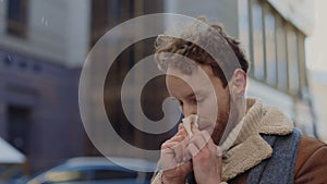 Young Caucasian man in winter clothes standing on the street and blows his nose into handkerchief. Symptoms of cold in