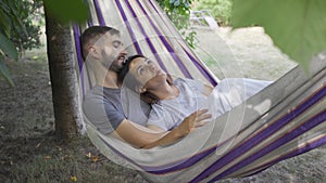 Young caucasian man and woman lying in hammock in the garden relaxing together. Loving couple together outdoors