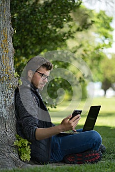 Young caucasian man working on his computer sitting under a tree while he is looking at his mobile at the same time. Technological