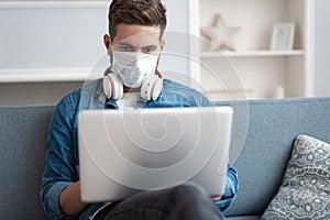 Young caucasian man working from home, wearing protective mask, using laptop. Coronavirus pandemic, covid 19 quarantine