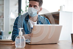Young caucasian man working from home, wearing protective mask, using laptop. Coronavirus pandemic, covid 19 quarantine